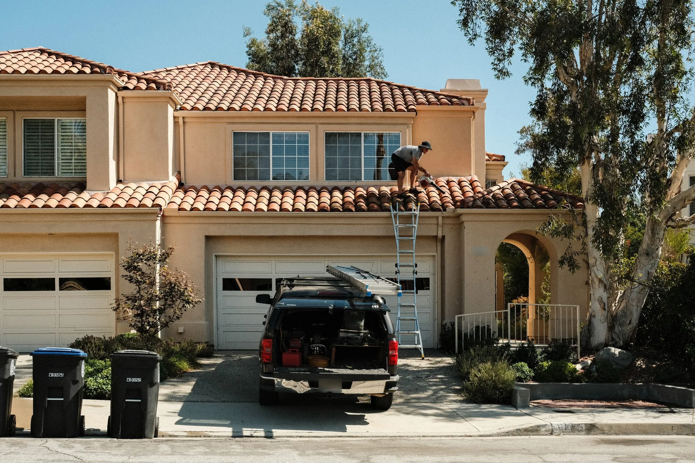 Squeegee Clean Windows truck in the driveway of a two-story tile-roof home while Elliot clears the gutters from the roof edge