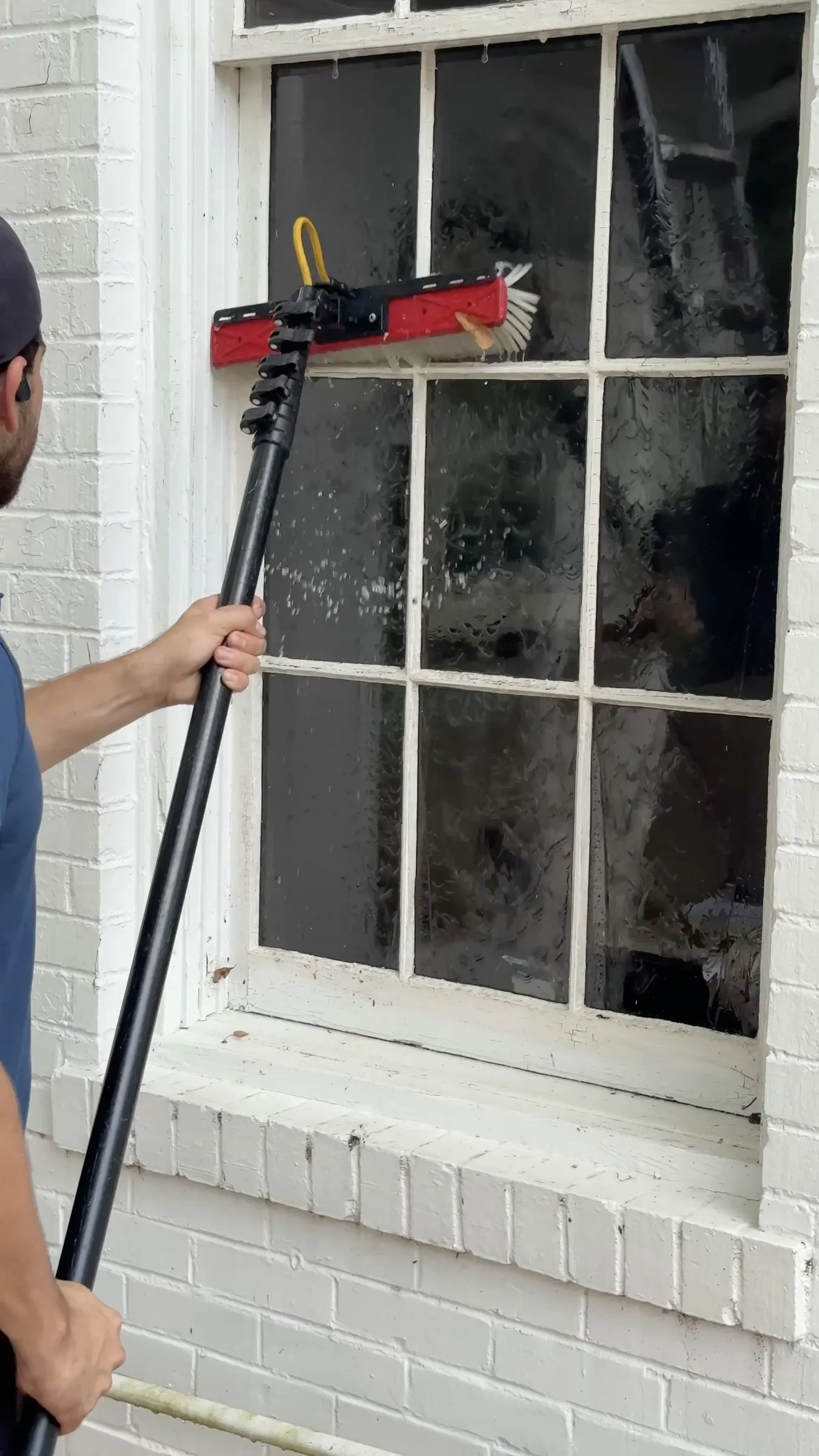 Water-fed brush scrubbing a multi-pane wood window on a painted brick facade